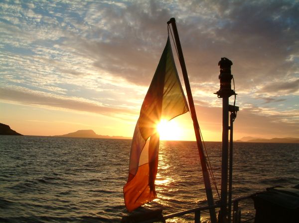 Amplitude by Gordan Kinsella. A Irish Naval vessel is at anchor and the sun is setting. The sunset is photographed through the ensign (the flag flying at a ships stern.) The suns light shines through the national flag of Ireland, and some coastal island are in the background in silhouette. The green, white, and orange are barely discernible. It is an image which stirs emotions of duty, service, and remembrance. A celestial body’s amplitude angle is the complement of its azimuth angle. when the body rises or sets, the amplitude angle is the arc of the horizon between the body and the East/West point of the horizon where the observer’s prime vertical intersects the horizon (at 90°). In practical navigation, a bearing of a body can be observed when it is on either the celestial or the visible horizon.