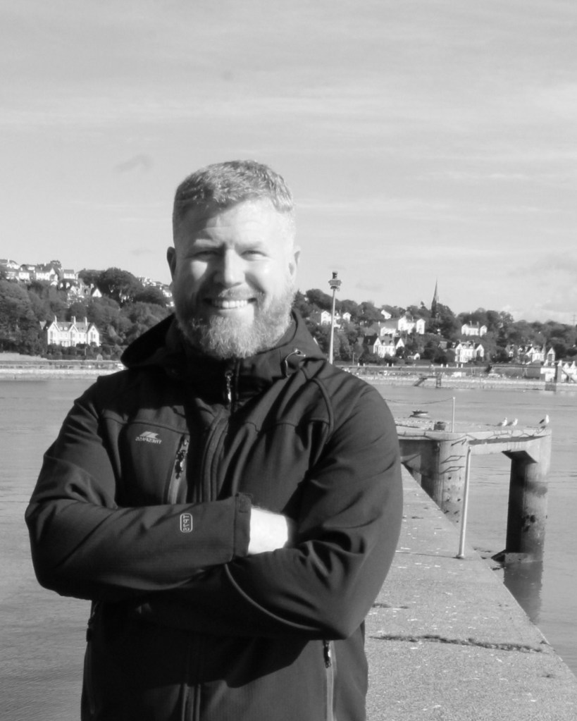 Ruairí de Barra smiling, arms folded, on a pier in Cobh harbour